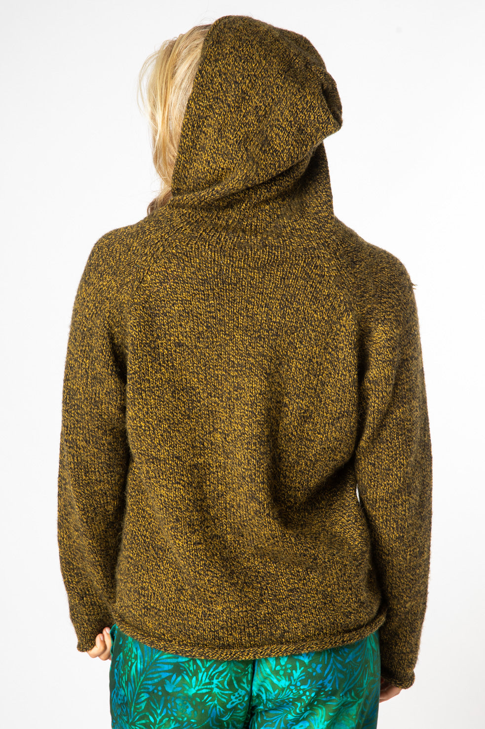 A blonde hippie girl facing backwards wearing a bumblebee alpaca hoodie with the hood up and green batik pants. 