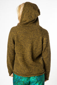 A blonde hippie girl facing backwards wearing a bumblebee alpaca hoodie with the hood up and green batik pants. 