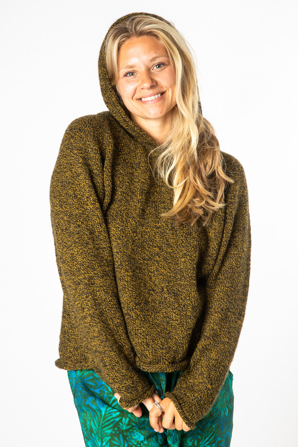 Smiling blonde hippie woman wearing a black and yellow women's alpaca sweater with the hood up and green patterned hippie bottoms. 