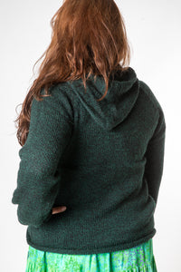 A brown haired hippie woman facing backwards wearing a dark green alpaca hoodie and a bright green patterned hippie skirt. 