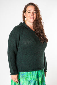 Beautiful hippie woman with brown curly hair wearing gold dangling earrings, a dark green alpaca hoodie, and a bright green patterned hippie skirt. 