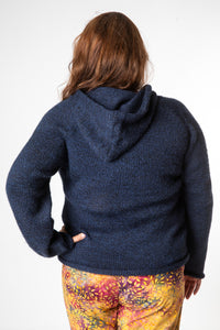 Backside of a brunette hippie woman wearing a women's alpaca hoodie in navy with yellow and red patterned hippie bottoms. 