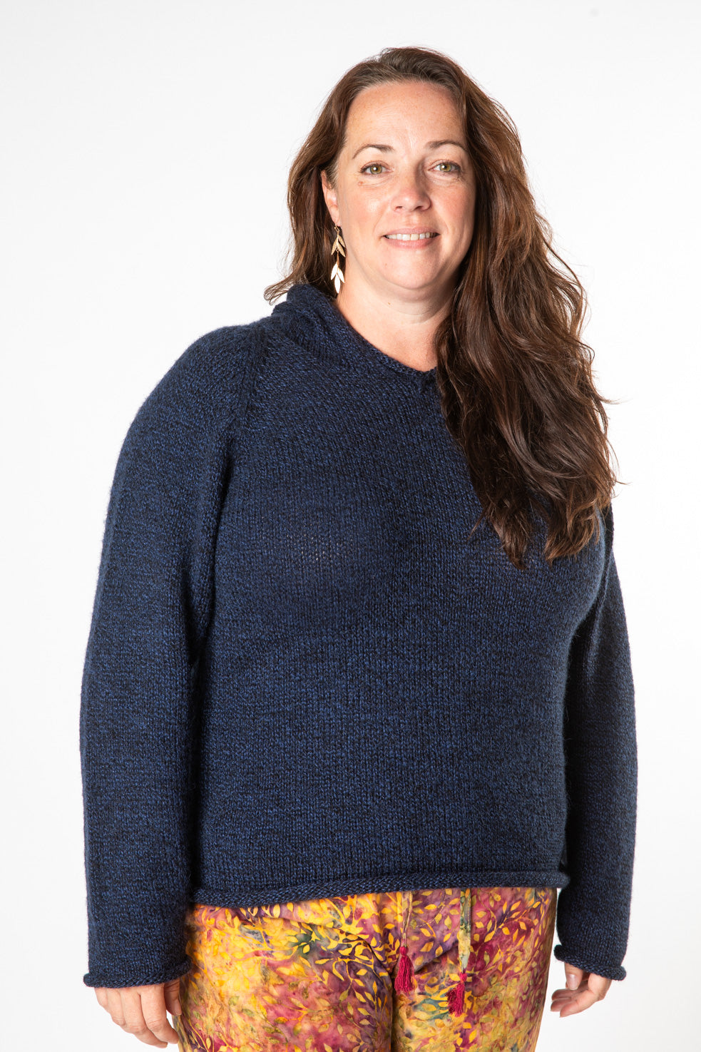 Happy bohemian woman with long brown curly hair wearing a navy women's alpaca sweater, gold dangling earrings, and yellow and red batik pants. 