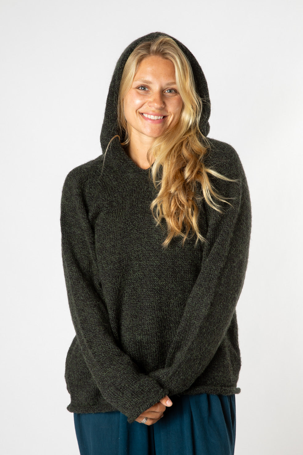 Happy smiling hippie girl with blonde wavy hair wearing a dark green women's alpaca hoodie with the hood up and blue hippie bottoms. 