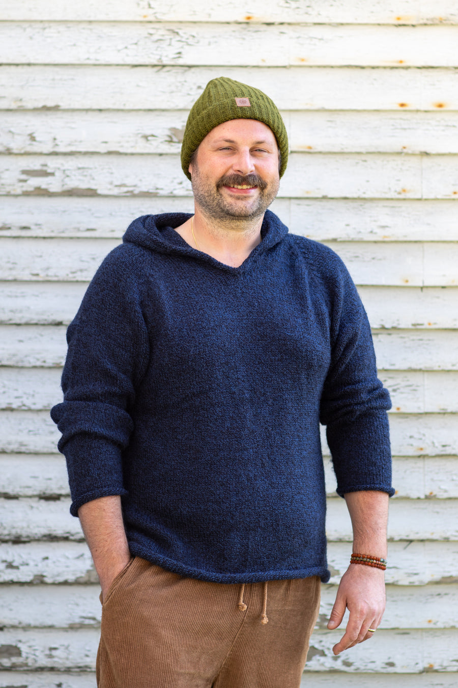 Handsome, rugged, mustache man smiling and wearing a hooded men's alpaca sweater with a green knit beanie and tan corduroy pants standing against a rustic white wall