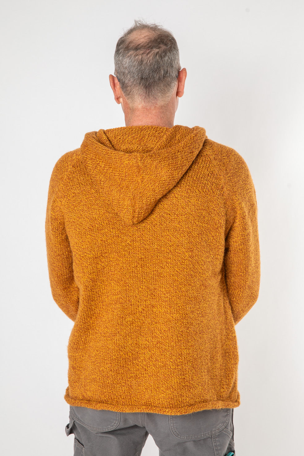 Backside of a hippie man with gray hair wearing a men's alpaca sweater with a hood in burnt orange with gray pants. 