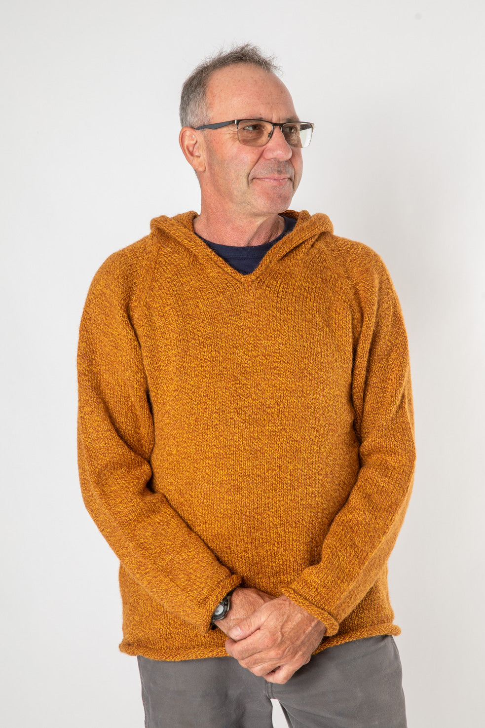 Handsome hippie man with gray hair and glasses looking into the distance waring a sunflower colored alpaca hoodie for men with gray hippie bottoms with his arms folded in front of him. 