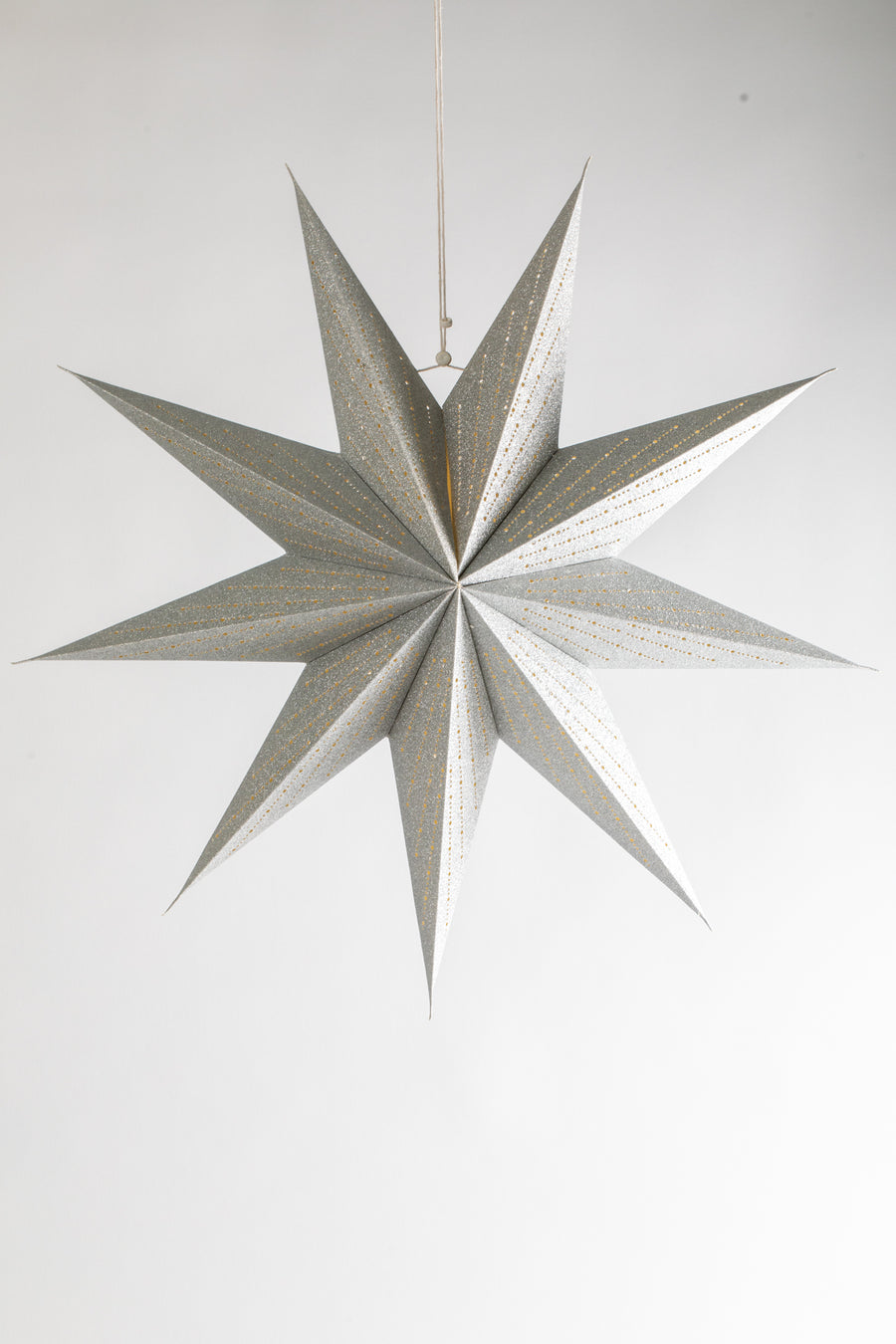 A glittering silver paper lantern in the shape of a 9-point star hanging in front of a white backdrop. 