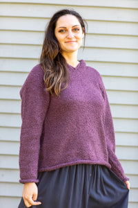 Beautiful brunette young woman wearing a hooded women's alpaca sweater in plum, with a navy bohemian skirt smiling while standing next to a white paneled wall. Classic hippie style photo