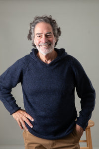 A smiling man with gray hair and a white beard standing in front of a stool wearing a navy alpaca sweater for men  and khaki hippie pants. 