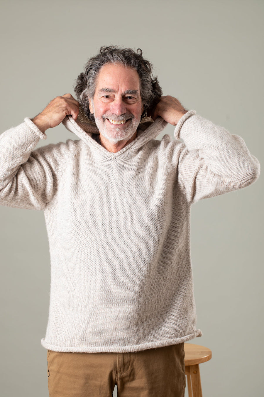 Handsome older hippie man with a white beard and gray hair standing in front of a stool moving to put on the hood of a white men's alpaca sweater. 