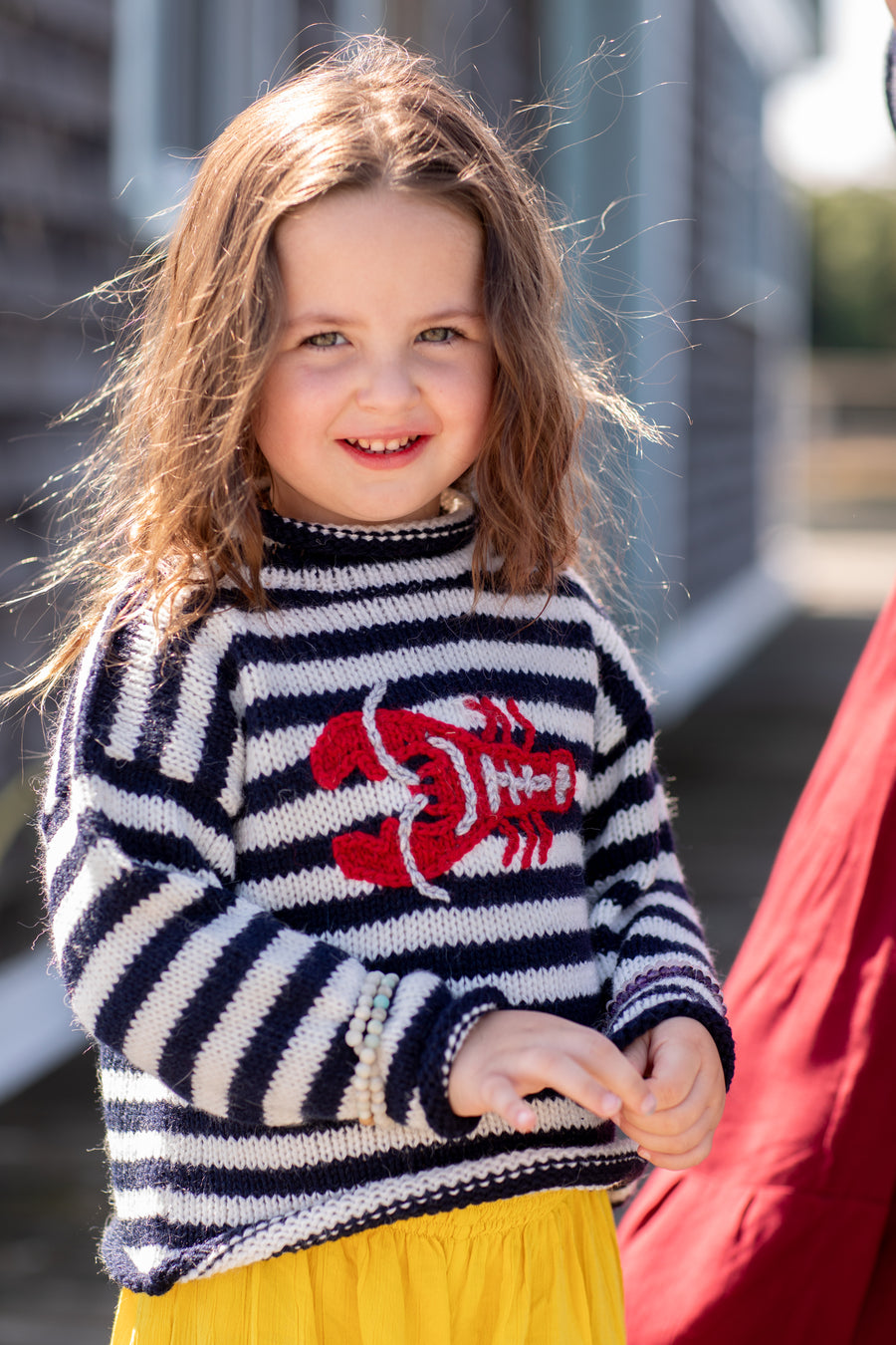 Adorable brunette little girl smiling at the camera wearing a blue and white nautical striped alpaca sweater with a red Maine lobster on the front, sitting in front of a rustic boathouse in Maine