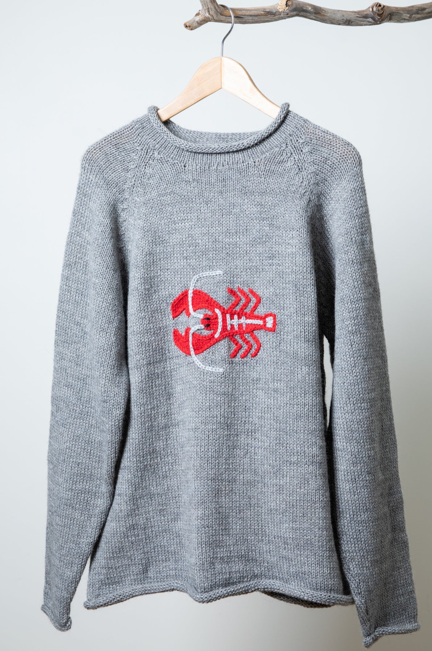 A gray lobster alpaca sweater hanging on a hanger from a tree branch featuring a red lobster decal. 