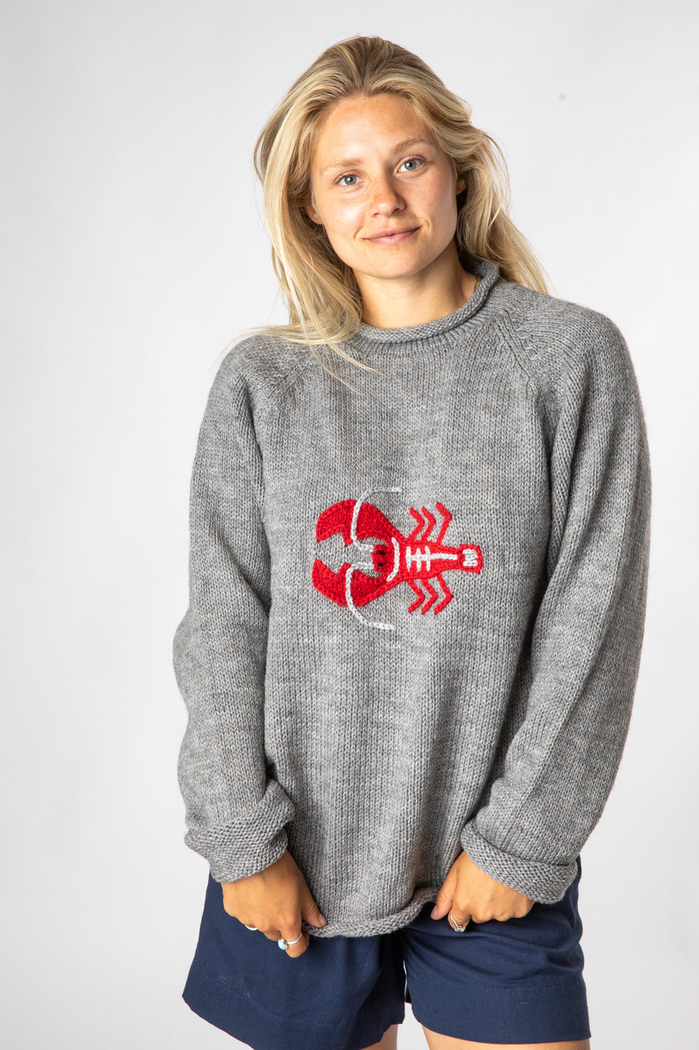 Beautiful blonde hippie girl smiling and wearing a gray lobster sweater made from alpaca wool and navy blue shorts. 