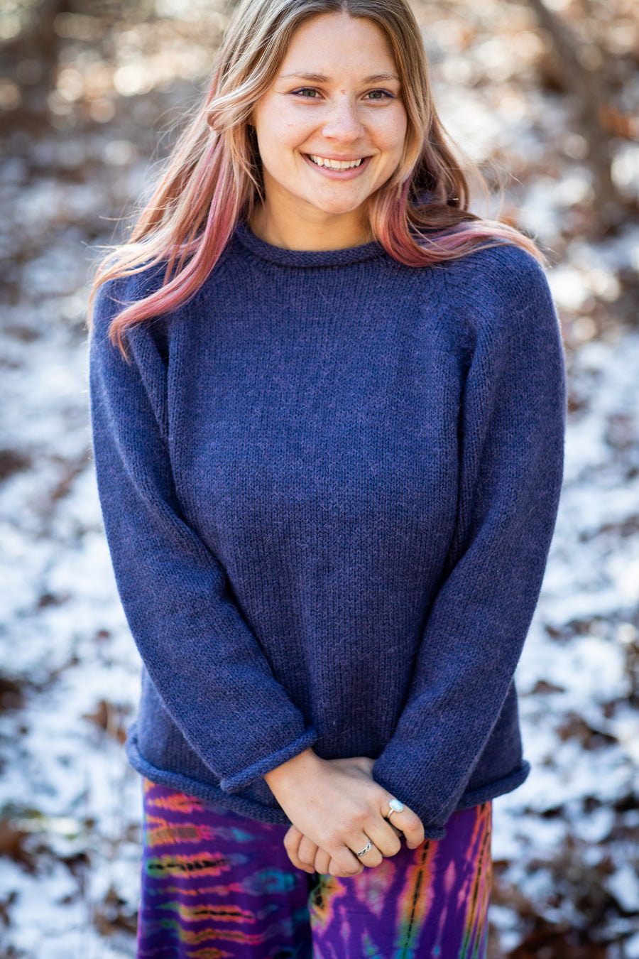 Beautiful smiling blonde hippie woman with pink hair wearing a roll neck women's alpaca sweater in Denim blue and tie dye pants standing in a snowy field