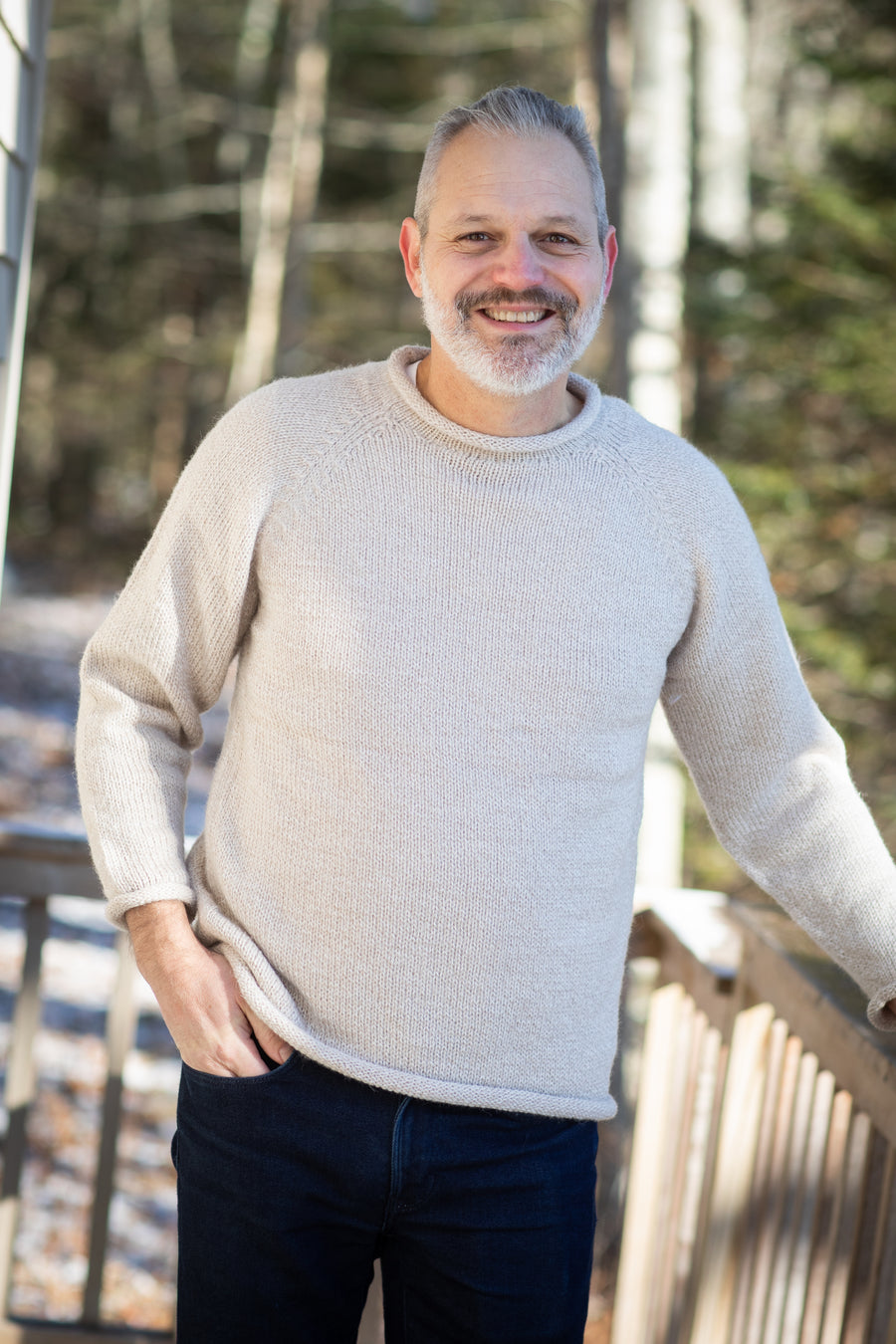 Handsome gray haired man wearing a mens alpaca sweater in Oatmeal and blue jeans, smiling and standing on a porch in the forest with pine trees in the background